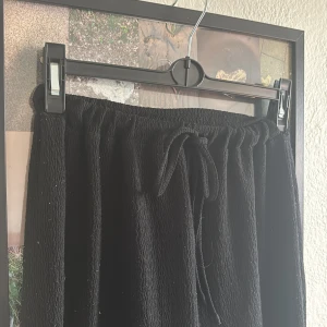 Svarta ribbade mjukisbyxor med snörning - Svarta mjukisbyxor med ribbad struktur och dragsko i midjan. Byxorna har en avslappnad passform och raka ben, perfekta för chill dagar. Materialet är mjukt och stretchigt för extra komfort.