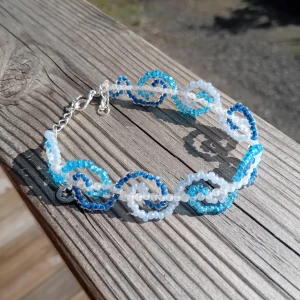 Blått och vitt pärlarmband med swirl - Handgjort unikt armband gjort av små pärlor i blått och vitt. Pärlorna är trädda i ett spiral/vågor mönster som ger en cool och lekfull look. Armbandet har justerbar kedja och karbinhake i silverfärg. Perfekt accessoar för att lyfta din outfit.