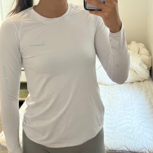 Vit långärmad Nike Dri-Fit träningströja - Säljer en vit långärmad träningströja från Nike med Dri-Fit-teknologi. Tröjan har rund hals, diskret Nike-logga på bröstet och texten 'Just Do It' på ärmen. Perfekt för löpning eller gym och sitter skönt mot kroppen.