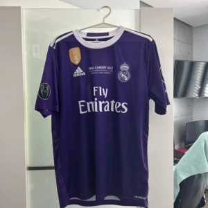 Real Madrid Ronaldo 7 Final Cardiff 2017 - Säljer en lila Real Madrid fotbollströja från Adidas, Final Cardiff 2017 edition med Ronaldo 7 på ryggen. Tröjan har vita detaljer, klubbmärke, Champions League-märken på ärmarna och Fly Emirates-tryck. Tillverkad i lätt och ventilerande material.