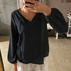 Mörkblå blus med v-ringning - Snygg mörkblå blus med v-ringning och tunna snören framtill. Blusen har långa ärmar med resår vid handlederna och ett luftigt, rakt fall. Perfekt för dig som gillar en stilren och enkel look.