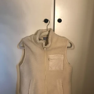 Beige fleeceväst från Lindex - Mysig beige fleeceväst från Lindex med hög krage och dragkedja framtill. Västen har en praktisk bröstficka med dragkedja i nylon och två sidofickor. Perfekt att slänga över en hoodie för extra värme.