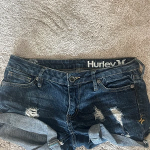 Mörkblå jeansshorts från Hurley - Snygga mörkblå jeansshorts från Hurley med slitna detaljer och upprullade benslut. Shortsen har klassiska fickor, nitar på ena bakfickan och coola slitningar framtill. Perfekta för sommardagar när du vill ha en avslappnad och trendig look. Säljs inte längre. midjemått är 35/36