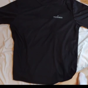 Svart Trussardi t-shirt med logga - Svart t-shirt från Trussardi med vit logga tryckt på bröstet och större logga på ryggen. Klassisk passform med rund hals och korta ärmar. Perfekt för dig som gillar stilrena och snygga basplagg med exklusiv känsla. Passar L XL