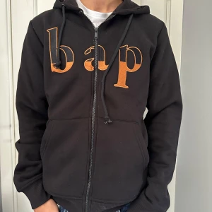Svart hoodie från Burgers & Pastrami - Svart hoodie med dragkedja och huva från Burgers & Pastrami. Framsidan har stora broderade bokstäver 'bap' i orange och guld, samt liten text nertill. Mjuk bomullskänsla och klassisk passform med fickor framtill.