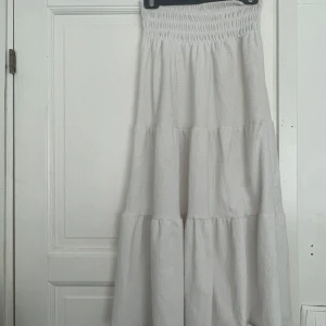 Vit långkjol från Gina Tricot - Supersöt vit långkjol från Gina Tricot med smock i midjan och volangiga lager. Kjolen är i ett lätt, strukturerat bomullstyg som känns perfekt för varma dagar. Enkel att styla med crop tops eller stickade tröjor för en chill vibe. Den är nopprig och har fläckar som jag inte vet om det kommer av därför säljer jag billigt 