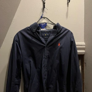 Mörkblå skjorta från Ralph Lauren - Klassisk mörkblå skjorta från Ralph Lauren med röd broderad logga på bröstet. Skjortan har lång ärm, knappar framtill och button-down krage. Classic fit-modell i mjuk bomull, perfekt för en clean och stilren look. Lite defekt i nacken fast syns ej när den är på