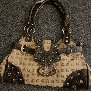Guess liknande handväska från Gamaya - Cool handväska från Gamaya i beige canvas med brunt mönster av G och solsymboler (väldigt likt märket Guess)Väskan har detaljer i mörkbrunt skinnimitation med croco-mönster, nitar, bälte och stor metallogo framtill. Flera fack med dragkedja och rejäla handtag. Perfekt för dig som gillar statement-accessoarer. OBS!!! den har fläckar nertill pga dricka som spillts (går att tvätta) när jag köpte den var spännena trasiga men det är inget man tänker på + handtagen är lite spretiga