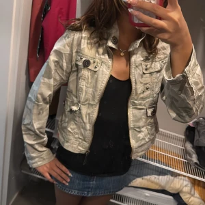 Beige camo bomberjacka med dragkedja - Säljer en beige och ljusgrön bomberjacka med camouflagemönster. Jackan har dragkedja framtill, flera fickor med knappar och coola broderade detaljer på ryggen. två knappar har tappats på framsidan. Det står att det är storlek M men passar mig i xs/s. 