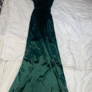 Mörkgrön långklänning i satin - Snygg mörkgrön långklänning i glansigt satinliknande material. Klänningen har en elegant oneshoulder-design med bred axelrem och figurnära passform upptill. Perfekt för bal eller festliga tillfällen när du vill sticka ut med en stilren look.