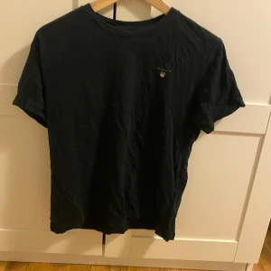 Svart t-shirt från GANT - En stilren svart t-shirt från GANT med liten broderad logga på bröstet. Klassisk rund halsringning och korta ärmar. Tillverkad i mjuk bomull som är skön att ha på sig hela dagen. Perfekt för en clean och enkel look.