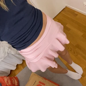 Ljusrosa volangkjol med shorts - Supersöt ljusrosa kortkjol med volangkant och inbyggda shorts under. Kjolen har en flowy känsla och bred linning som sitter snyggt i midjan. Perfekt för dig som vill ha en gullig och bekväm look till sommaren.