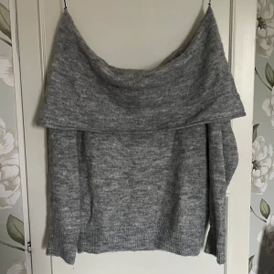 Grå offshoulder stickad tröja - Mysig grå stickad tröja med bred offshoulder-krage som ger en avslappnad och trendig look. Perfekt för kyliga dagar och enkel att styla med jeans eller kjol. Tröjan har ribbade muddar och en lös passform för extra komfort.