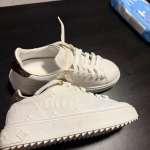 Louis Vuitton vita sneakers med logga - Exklusiva vita sneakers från Louis Vuitton med präglad LV-logga på sidan och brunt parti med guldigt LV-märke på hälen. Skorna har kraftig platt sula med mönster och detaljer i skinn. Snygga guldfärgade öljetter och klassisk snörning.