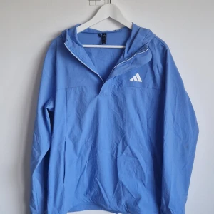 Blå vindjacka från Adidas med huva - Blå vind/vattenavvisande jacka från Adidas med vit logga på bröstet. Jackan har huva, halv dragkedja. Har en väldigt liten fläck/prick på ärmen (se sista bilden), annars är den som ny