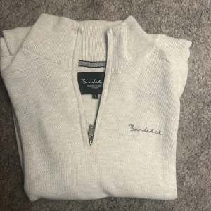 Vit half zip tröja från Bondelid - Snygg vit långärmad tröja från Bondelid med half zip och hög krage. Tröjan är stickad i bomull och har diskret broderad logga på bröstet. Perfekt för lager-på-lager och chill stil.