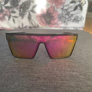 Svarta furturistiska solglasögon med spegelglas - Säljer ett par feta solglasögon med svart båge och stora, spegelblanka glas i rosa och gult. Glasögonen har en raggig vibe med rektangulär form och bred front. Perfekta för dig som vill sticka ut och få en cool look i sommar.