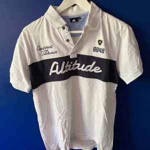 Vit pikétröja från 8848 Altitude M - Vit pikétröja från 8848 Altitude med svart horisontellt band och broderad text 'Altitude' över bröstet. Klassisk krage med knappar och broderade detaljer på framsidan. Perfekt för en sportig och stilren look.