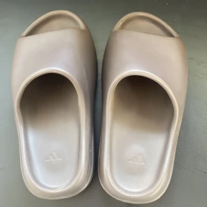 Adidas Yeezy Slide i brun - Säljer ett par Adidas Yeezy Slide i beige färg. Slidesen har en chunky, ribbad sula och minimalistisk design med öppen tå. Tillverkade i syntetmaterial för en mjuk och bekväm känsla. Perfekta för chill dagar och streetwear-stil.
