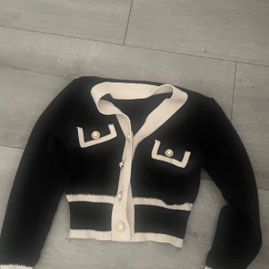 Svart och beige kofta med pärlknappar - Snygg svart kofta med beige detaljer och pärlknappar. Croppad modell med långa ärmar, dekorativa fickor framtill och ribbade kanter. Perfekt för dig som gillar en chic och trendig look.