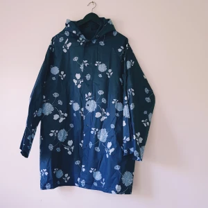 Blommig regnjacka i mörkblått L/XL - Snygg mörkblå regnjacka med stora blommiga mönster i vitt. Jackan har huva, långa ärmar och är tillverkad i 100% EVA-material som står emot regn. Perfekt för att hålla sig torr och ändå sticka ut med cool stil.  Dock små hål bak (se extra bilder)