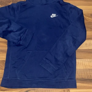 Mörkblå Nike hoodie XL 158-170 - Säljer en mörkblå hoodie från Nike i storlek XL. 158-170 Tröjan har klassisk huva, magficka och Nike-logga broderad på bröstet. Tillverkad i mjuk bomullsmix och har långa ärmar med ribbade muddar. Perfekt för chill dagar eller när du vill ha en sportig look.