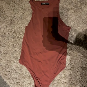 Rostbrun ärmlös bodysuit från SHEIN - Snygg rostbrun bodysuit från SHEIN med hög hals och ärmlös design. Tight passform som framhäver siluetten och passar perfekt till både jeans och kjol. Gjord i mjukt, stretchigt syntetmaterial för en bekväm känsla hela dagen.