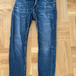 Blå jeans från Tiger of Sweden, 29/30 - Snygga blå jeans från Tiger of Sweden i klassisk tvätt med diskreta slitningar. Modellen har raka ben och normal passform, med orangea sömmar och femficksdesign. Perfekta för dig som gillar stilrena och tidlösa jeans.