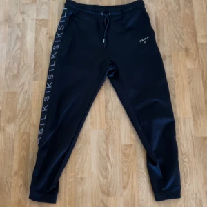 Svarta mjukisbyxor från SikSilk XL - Svarta mjukisbyxor från SikSilk i storlek XL. Byxorna har snörning i midjan och tryckt logga längs ena benet. De är tillverkade i mjukt material och har en slim passform med muddar vid bensluten. 