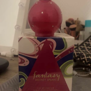 Fantasy Britney Spears parfym 100ml - Fantasy av Britney Spears är en ikonisk parfym i en rund, mörkrosa flaska med små glittriga stenar och dekorativ hals. Kommer i en färgglad kartong med rosa, gröna och blå mönster. Eau de Parfum, 100 ml spray. Perfekt för dig som gillar söta och fruktiga dofter.