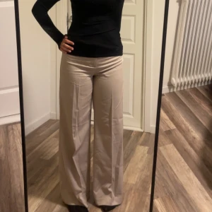 Beiga vida kostymbyxor - Säljer ett par snygga beiga kostymbyxor med hög midja och riktigt breda ben. Byxorna har pressveck framtill och är tillverkade i ett mjukt material som känns skönt mot huden. Perfekta för dig som gillar en stilren och trendig look.