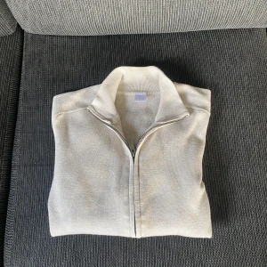 Beige stickad kofta från Lindex - Snygg beige stickad kofta från Lindex med hel dragkedja framtill och ribbade muddar. Perfekt för lager-på-lager och funkar till både jeans och t-shirt. Enkel och stilren design som passar till många olika looks.