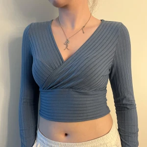 Blå ribbad omlottopp croppad - Säljer en blå croppad omlottopp med långa ärmar. Toppen är ribbad och har en snygg v-ringning framtill. Perfekt för dig som gillar en stilren men trendig look. Passar till både jeans och kjol för en cool outfit. Finns även i vit och svart!