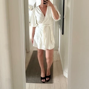 Vit omlottklänning med puffärm - Superfin vit omlottklänning från Wakakuu icons med puffiga korta ärmar och knyt i midjan. Klänningen är kort och har en v-ringad halsringning som ger en snygg siluett. Perfekt för sommarens alla tillfällen!