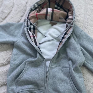 Grå hoodiejacka från Burberry - Grå hoodiejacka från Burberry med klassiskt rutigt foder i huvan. Jackan har dragkedja, snörning i huvan och stora fickor framtill. Tillverkad i mjuk bomullsmix, perfekt för chill dagar. Ikoniska Burberry-detaljer på snörstopparna och etiketten.