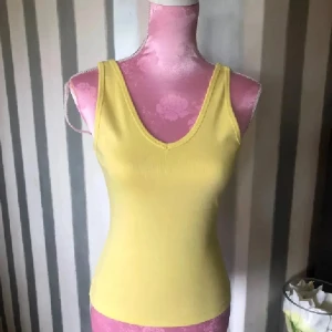 Gul ribbad linnetopp med v-ringning - Snygg gul linnetopp med v-ringning både fram och bak. Toppen är ribbad och har en tight passform som framhäver siluetten. Perfekt för varma dagar och enkel att matcha med jeans eller kjol.
