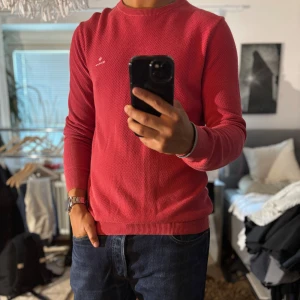 Röd stickad tröja från GANT - Säljer en snygg röd stickad tröja från GANT med diskret logga på bröstet. Tröjan har rund halsringning, ribbade muddar och är långärmad. Perfekt för dig som gillar klassisk och stilren look med en pop av färg.