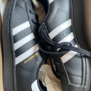 Adidas Superstar svartvita sneakers - Klassiska Adidas Superstar sneakers i svart skinn med vita ränder och svart sula. Ikonisk design med snörning och guldfärgad Superstar-text på sidan. Perfekta för dig som gillar streetstyle och tidlös look.