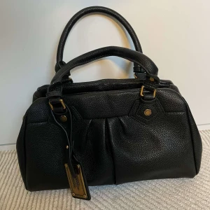 Svart handväska från Marc by Marc Jacobs - Snygg svart handväska i skinn från Marc by Marc Jacobs med två handtag, guldfärgade metalldetaljer och dragkedja upptill. Väskan har en klassisk form och en tag med logga framtill. Perfekt för dig som gillar stilrena accessoarer.