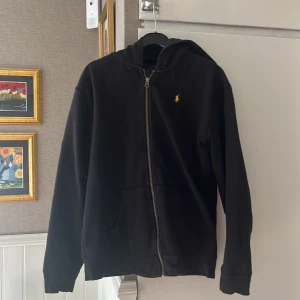 Svart hoodie från Polo Ralph Lauren - Svart hoodie från Polo Ralph Lauren med dragkedja och klassisk gul broderad logga på bröstet. Tröjan har huva, två fickor fram och är tillverkad i mjuk bomull. Perfekt för en chill och stilren look.