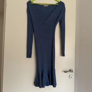 Blå ribbad långklänning Vanille & Chocolat - Supersnygg blå långklänning från Vanille & Chocolat med ribbad struktur, v-ringad omlott upptill och långa ärmar. Klänningen har en volangkant nertill som ger extra rörelse och en feminin touch. Perfekt för dig som gillar en tight och stilren look.