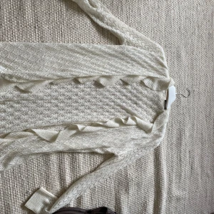 Vit stickad kofta med volangkant - Superfin vit stickad kofta med öppen front och dekorativ volangkant längs hela öppningen. Koftan har ett genombrutet mönster och långa ärmar med ribbade muddar. Inga skavanker eller defekter❤️