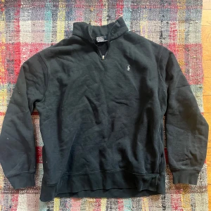 Svart tröja med dragkedja från Polo Ralph Lauren - Säljer en stilren svart tröja från Polo Ralph Lauren med halv dragkedja och broderad logga på bröstet. Perfekt för en avslappnad look. Tröjan har långa ärmar och är gjord i ett mjukt material.