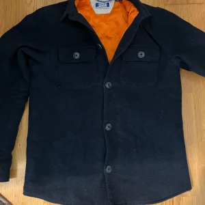 Blå overshort från Jack & Jones - Snygg svart overshirt från Jack & Jones med orange quiltat foder. Jackan har knappar framtill och två bröstfickor med knappar. Perfekt för en stilren look.