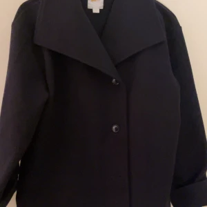 Svart kappa med knappar Gina trickot  - Helt ny använd 1 gång Elegant svart kappa med stora slag och knäppning framtill. Perfekt för kyligare dagar med sin stilrena design och långa ärmar. Från Gina 