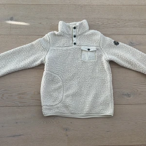 Vit fleece tröja från Pelle P - Mysig vit fleece tröja från Pelle P med hög krage och knappar framtill. Tröjan har en praktisk bröstficka med knapp och en rund logga på ärmen. Perfekt för kyliga dagar!