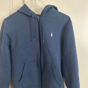 Blå hoodie från Ralph Lauren - Säljer en snygg blå hoodie från Ralph Lauren med dragkedja och klassisk logga på bröstet. Den har en justerbar huva med snören och praktiska fickor framtill. Perfekt för en avslappnad stil.