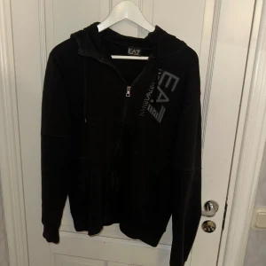 Svarta mjukisdress från Emporio Armani - Snygg svart dress från Emporio Armani med EA7-logga på ben o bröst. Perfekta för en avslappnad stil. Byxorna har en elastisk midja för extra komfort.