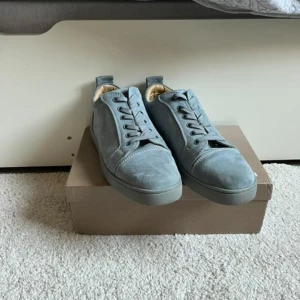 Grå sneakers från Christian Louboutin - Snygga grå sneakers i mocka från Christian Louboutin med den ikoniska röda sulan. Skorna har snörning och en rund tå, vilket ger en stilren look. Perfekta för dig som vill ha en touch av lyx i vardagen.