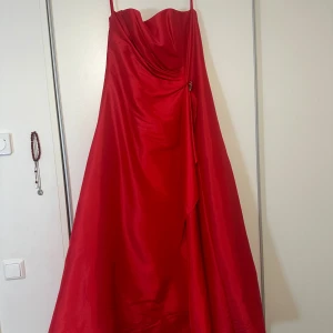 Röd långklänning med dekorativ detalj - Elegant röd långklänning utan axlar och en vacker drapering framtill. Klänningen har en dekorativ broschdetalj och knappar längs ryggen. Perfekt för speciella tillfällen. Storlek L-XL